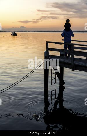 Vater gibt Sohn Huckepack Fahrt auf einem hölzernen Pier am Meer während schönen Sonnenuntergang Stockfoto
