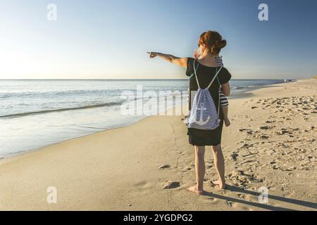 Junge Frau am Strand hält ein Kleinkind in ihrem Arm und zeigt auf den Ozean Stockfoto