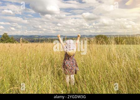 Nettes kleines blondes Mädchen, das im Sommer auf einem Feld steht und ihre Arme voller Freude hebt Stockfoto