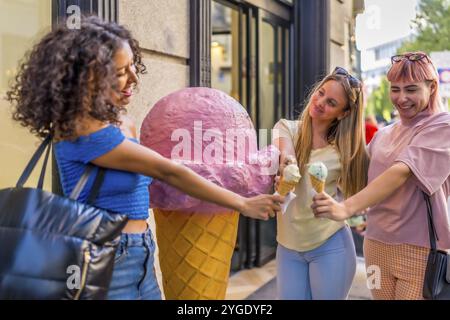 Verschiedene Frauen, die Spaß haben, Eis zu essen, neben riesigen Eiskegel, die den Eingang eines Ladens schmücken Stockfoto