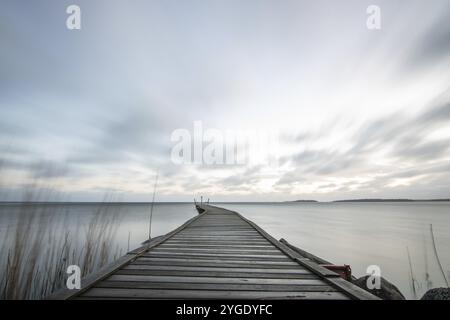 Sonnenaufgang über einem See im Herbst. Ein Holzsteg ragt ins Wasser, während sich Gras am Sandstrand angesiedelt hat. Der See und der Strand am V-See Stockfoto
