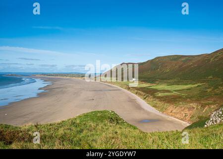 Blick hinunter auf Rhossili Beach auf der Gower Halbinsel von einem Hügel und zeigt einen großen einsamen Sandstrand mit bunten Hügeln unter blauem Himmel Stockfoto