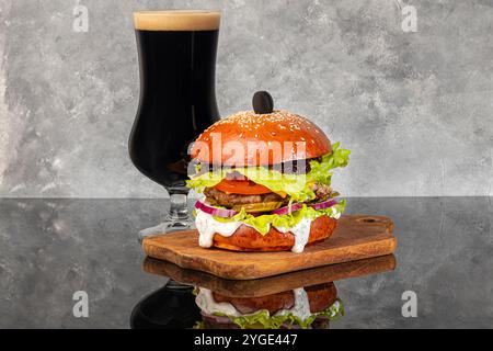 Burger auf einem Holzbrett mit einem Patty, Tomate, Käse, Gurke und Salat. Ein Glas Bier im Hintergrund. Kopierbereich Stockfoto