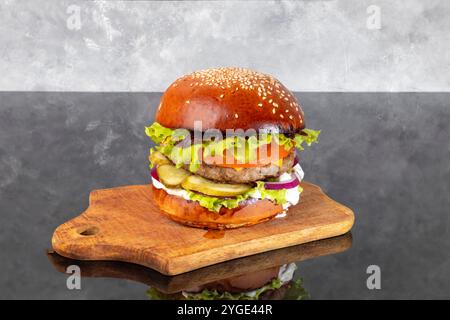 Burger auf einem Holzbrett mit einem Patty, Tomate, Käse, Gurke und Salat. Ein Glas Bier im Hintergrund. Kopierbereich Stockfoto