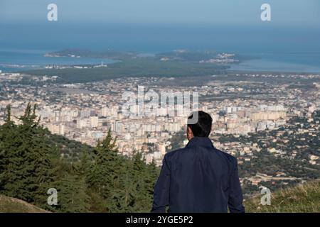 Junger Mann, der auf einer Klippe in den Bergen steht und den Blick auf die Stadt Vlore in Albanien genießt. Ein Mann, der auf die Stadt im Tal blickt und auf der Th steht Stockfoto