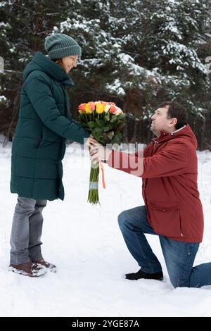 Verliebtes Paar im Winter im Park. Der Mann geht auf ein Knie und schenkt seiner geliebten Frau einen großen Strauß Rosen. Valentinstag. Romantische Beziehungen Stockfoto
