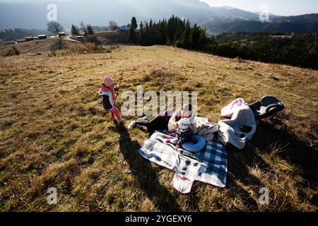 Eine Mutter und ihre Kinder machen ein Picknick auf einem grasbewachsenen Hügel, umgeben von malerischen Bergen. Sonniges Wetter und Freizeit im Freien sorgen für ein warmes familienatmos Stockfoto