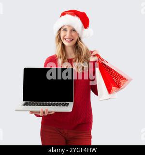Weihnachtseinkäufe im Internet, lächelnde Frau im weihnachtsmann-Hut zeigt Laptop-Computer leeren Bildschirm, während sie eine Geschenktüte hält, Adv für Top-Angebote zu Weihnachten Stockfoto