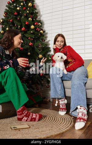 Zwei Freunde feiern gemeinsam die Weihnachtszeit und kuscheln sich mit ihrem flauschigen Bichon Frise am Weihnachtsbaum. Stockfoto