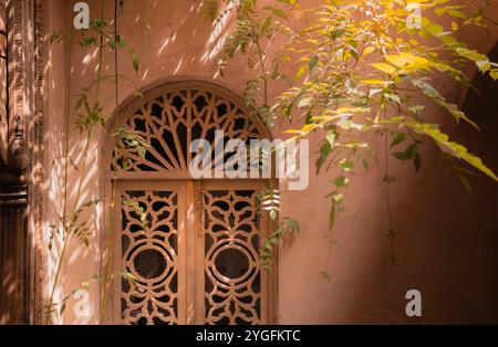 Kunstvolle marokkanische traditionelle Holztür mit kompliziertem Gitterdesign, strukturierter Wand und umliegendem Grün in einem ruhigen, luxuriösen Riad-Innenhof mit Stockfoto