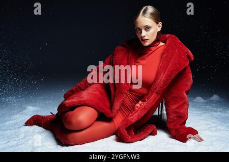 Eine atemberaubende junge Frau sitzt elegant auf dem Schnee und zeigt ihre leuchtend rote Kleidung mit Pose. Stockfoto