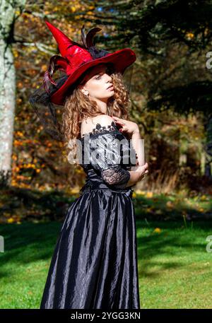 Eine Hexe in einem langen schwarzen Kleid und einem Hexenhut im Park draußen in der rauen Herbstsonne in Schottland Stockfoto