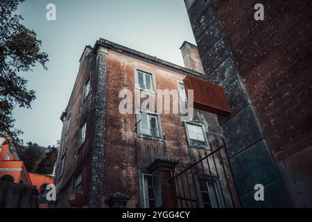 Weitwinkelaufnahme eines alten, verwitterten Backsteingebäudes mit verblasster Fassade und gerahmten Fenstern. Ein rostiges Modellschild ragt aus der Wand hervor und fügt ein V hinzu Stockfoto