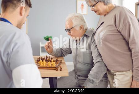 Junger Schwesternmann beim Schachspiel mit einem grauhaarigen älteren Ehepaar in der Klinik. Stockfoto