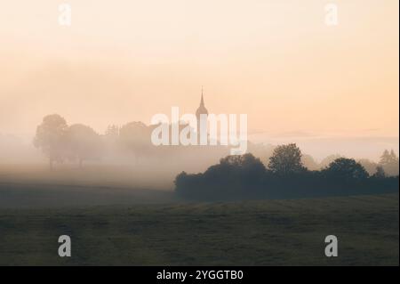 Blick über Wiesen und Hecken zu einem Kirchturm im Nebel im Morgenlicht am Ammersee in Bayern Stockfoto