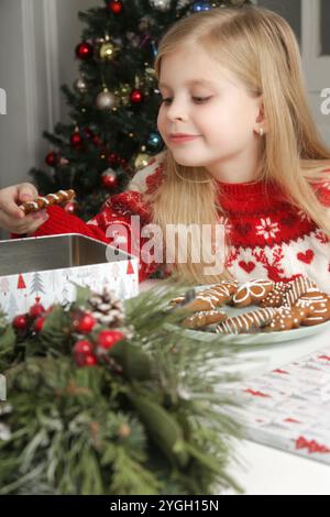 Entzückendes kleines Mädchen, das zu Hause Winterferien genießt, Weihnachtspullover trägt, Lebkuchenkekse auf dem Tisch, gemütliche festliche Atmosphäre. Stockfoto