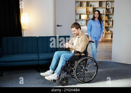 Ein süßer junger Mann im Rollstuhl hält ein Handy, während seine Freundin ihm hilft, sich in der Wohnung zu bewegen. Stockfoto