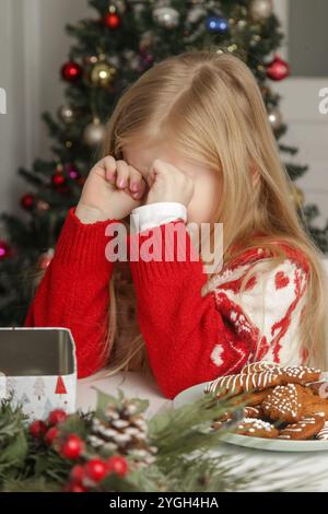 Trauriges, frustriertes kleines Mädchen im Weihnachtsoutfit und Weihnachtsmann-Hut am Tisch mit Lebkuchenkeksen, Winterurlaub-Stress-Konzept. Stockfoto