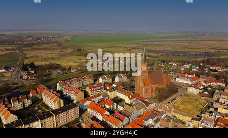 Drohnenblick auf die Trzebiatów gotische Kirche aus rotem Backstein und ihren Turm, der über den Gebäuden der Stadt ragt, umgeben von Feldern unter einem wolkenbedeckten Himmel. Stockfoto