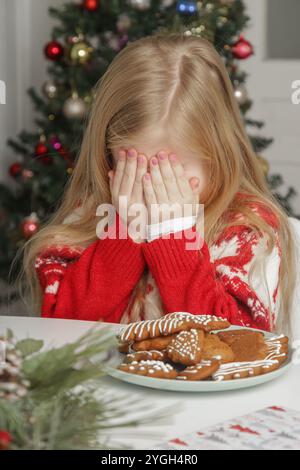 Trauriges, frustriertes kleines Mädchen im Weihnachtsoutfit und Weihnachtsmann-Hut am Tisch mit Lebkuchenkeksen, Winterurlaub-Stress-Konzept. Stockfoto