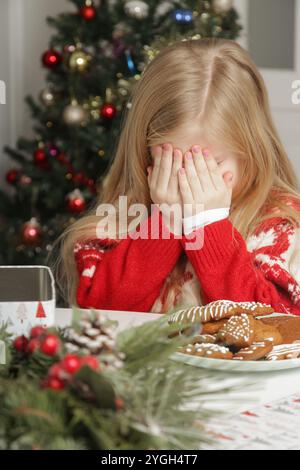 Trauriges, frustriertes kleines Mädchen im Weihnachtsoutfit und Weihnachtsmann-Hut am Tisch mit Lebkuchenkeksen, Winterurlaub-Stress-Konzept. Stockfoto