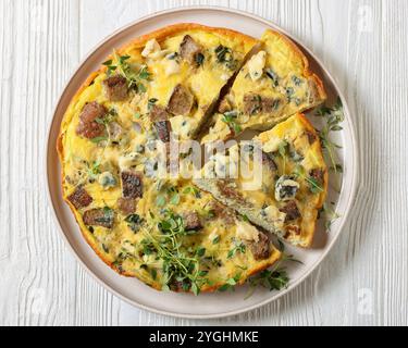 Gebackenes Omelette mit Blauschimmelkäse, Roggencroutons und frischem Thymian auf Teller auf weißem Holztisch, horizontaler Blick von oben, flache Lage, Nahaufnahme Stockfoto