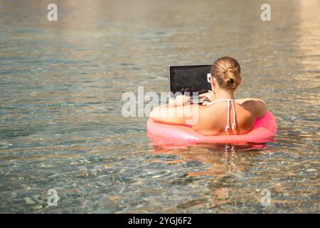 Eine Frau sitzt auf einem pinkfarbenen Schlauchboot im Wasser und benutzt einen Laptop. Das Konzept der Entspannung und Freizeit, während die Frau ihre Zeit am genießt Stockfoto