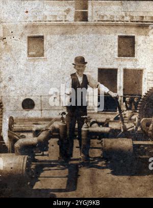 Dieses Vintage-Foto zeigt einen Mann in Kleidung aus dem 19. Jahrhundert, einschließlich eines Bowler-Hutes, der neben großen Zahnrädern und Rohren auf dem Deck eines Dampfschiffes steht. Die maritime Umgebung und seine Kleidung deuten auf einen Zeitrahmen der späten 1800er bis frühen 1900er Jahre hin, wahrscheinlich während der Spitzenzeiten dampfbetriebener Schiffe. Das Bild bietet einen seltenen Einblick in die historische Seefahrertechnik und das Leben auf Schiffen und bewahrt ein Stück der Industrie- und maritimen Geschichte dieser Epoche. Stockfoto