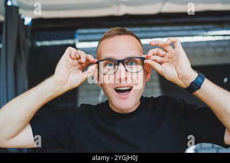 Ein süßer junger Mann in Brille und schwarzem T-Shirt steht auf einer Sommerterrasse in einem Café und lächelt in die Kamera. Remote-Sommerjob online. Herren Stockfoto