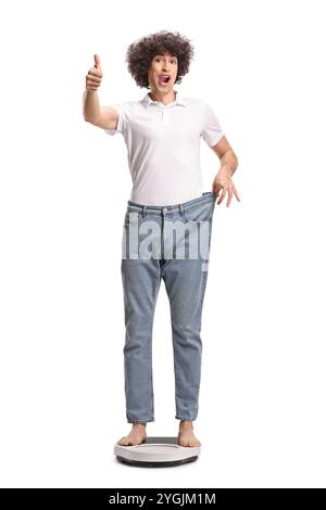 Junger Mann auf einer Waage, der eine große Jeans trägt und die Daumen auf weißem Hintergrund nach oben bewegt Stockfoto