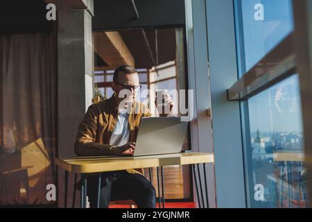 Ein ausgewogener Mann in Brille und lässiger Kleidung arbeitet an einem Laptop von einem gemütlichen Arbeitsplatz aus. Ein erfolgreicher Freelancer arbeitet Remote. Stockfoto