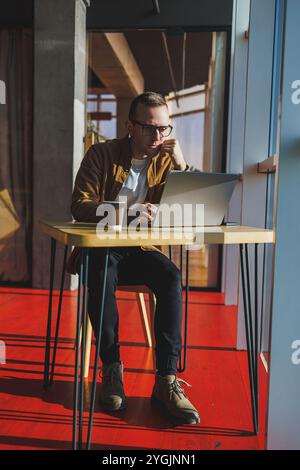Ein ausgewogener Mann in Brille und lässiger Kleidung arbeitet an einem Laptop von einem gemütlichen Arbeitsplatz aus. Ein erfolgreicher Freelancer arbeitet Remote. Stockfoto