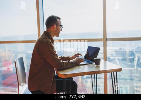 Ein ausgewogener Mann in Brille und lässiger Kleidung arbeitet an einem Laptop von einem gemütlichen Arbeitsplatz aus. Ein erfolgreicher Freelancer arbeitet Remote. Stockfoto