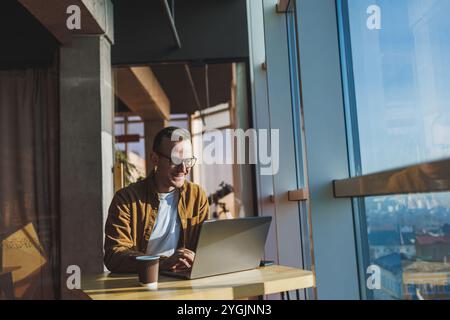 Ein ausgewogener Mann in Brille und lässiger Kleidung arbeitet an einem Laptop von einem gemütlichen Arbeitsplatz aus. Ein erfolgreicher Freelancer arbeitet Remote. Stockfoto