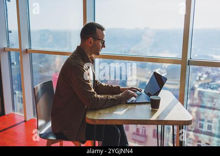 Ein ausgewogener Mann in Brille und lässiger Kleidung arbeitet an einem Laptop von einem gemütlichen Arbeitsplatz aus. Ein erfolgreicher Freelancer arbeitet Remote. Stockfoto