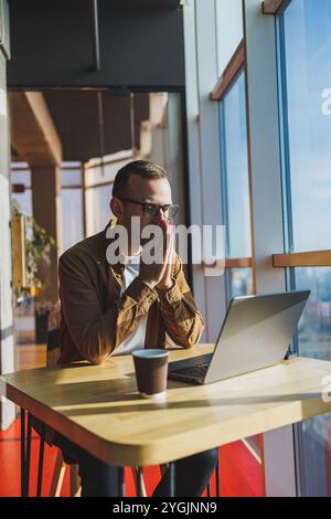 Ein ausgewogener Mann in Brille und lässiger Kleidung arbeitet an einem Laptop von einem gemütlichen Arbeitsplatz aus. Ein erfolgreicher Freelancer arbeitet Remote. Stockfoto