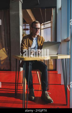 Ein ausgewogener Mann in Brille und lässiger Kleidung arbeitet an einem Laptop von einem gemütlichen Arbeitsplatz aus. Ein erfolgreicher Freelancer arbeitet Remote. Stockfoto