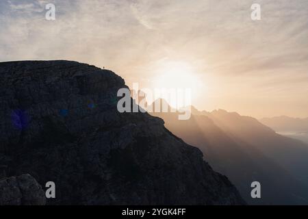 Die Silhouette eines Wanderers steht auf einem felsigen Abgrund, am Horizont taucht die Sonne die Landschaft in goldenes Licht. Stockfoto