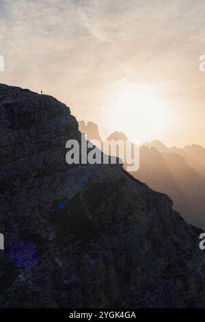 Die Silhouette eines Wanderers steht auf einem felsigen Abgrund, am Horizont taucht die Sonne die Landschaft in goldenes Licht. Stockfoto