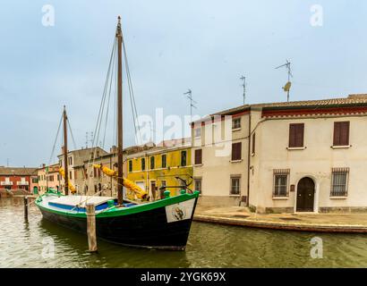 Comacchio, Ferrara – IT – 16. Oktober 2024 Ein kleines, vertäutes Boot auf einem schmalen Kanal, der von pastellfarbenen Gebäuden gesäumt wird. Der nasse Kopfsteinpflastergang verleiht Charme auf einem M Stockfoto