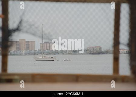 POV-führende Linien, Blick durch Maschendrahtzaun auf beschädigtem Holzsteg. Blick auf ein Segelboot in Boca Ciega Bay Gulfport Florida Beach. Nach St Stockfoto