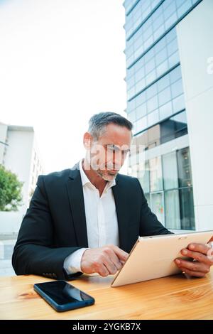 Vertikal. Geschäftsmann mit Tablet in einem Café im Freien erfolgreicher professioneller Manager mittleren Alters, der während der Kaffeepause in der Stadt mit einem digitalen Touchpad arbeitet. Hochwertige Fotos Stockfoto