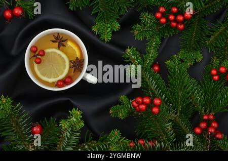 Tasse mit Getränk und Zitrone, Orange, Anissternen, Zimtstangen und weihnachtlichen roten Dekorationen, grünen Zweigen auf dunklem Hintergrund. Stockfoto