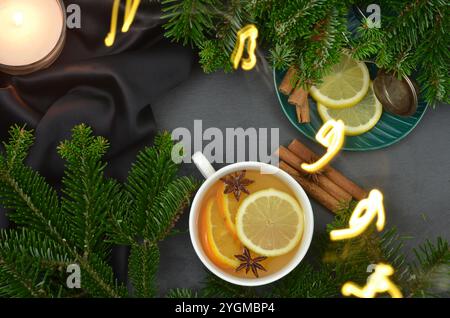 Brennende Kerze und Weihnachtslichter, Tasse mit Getränk, Zitrone, Anissterne, Zimtstangen, dunkler Hintergrund. Stockfoto