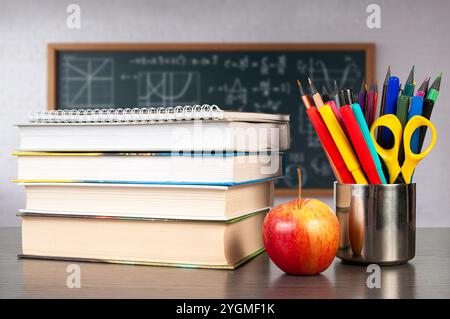 Ein Stapel Lehrbücher befindet sich auf einem Schreibtisch, neben einem hellroten Apfel und einem Behälter mit farbenfrohen Schreibwaren, ideal für eine Unterrichtsstunde. Stockfoto