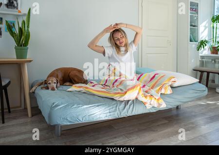 Gerade aufgewachte Frau sitzt im Bett und dehnt sich mit erhobenen Armen und geschlossenen Augen, genießt Ruhe. Stockfoto