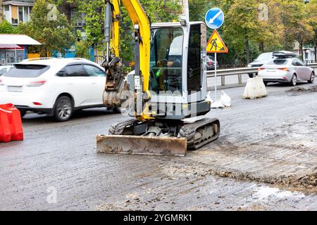 Das Baupersonal verwendet einen kompakten Bagger, um an einem regnerischen Tag eine stark befahrene Stadtstraße zu reparieren Stockfoto