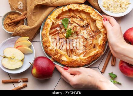 Eine Person legt vorsichtig eine frisch gebackene Apfelgalette auf einen Tisch, umgeben von geschnittenen Äpfeln, Zimtstangen und Mandelflocken. Die warme Küche Stockfoto