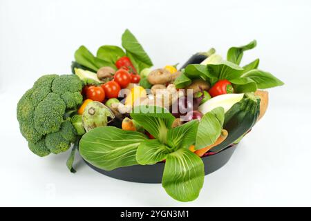 Rohes gemischtes Bio-Gemüse in einer dunklen Schüssel auf weißem Hintergrund. Einschließlich Brokkoli, Pac Choi, rote Zwiebeln, Knoblauch, Rosenkohl, Tomaten Stockfoto