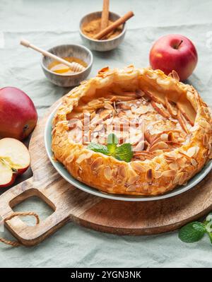 Eine frisch gebackene Apfeltorte sitzt auf einem rustikalen Holzbrett, garniert mit Mandeln und Minze, begleitet von roten Äpfeln und einer kleinen Schüssel Honig, Inviti Stockfoto